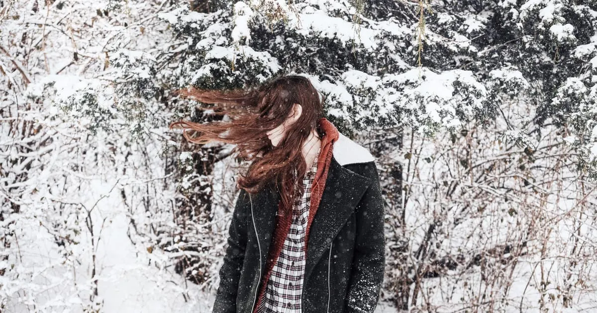 Femme aux longs cheveux châtains soulevés par le vent, en manteau d'hiver, dans une forêt enneigée