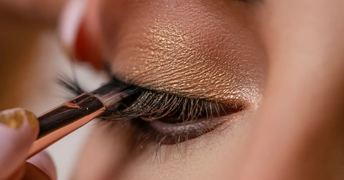 Gros plan d'un œil maquillé d'un fard doré scintillant, un pinceau déposant le produit au ras des cils