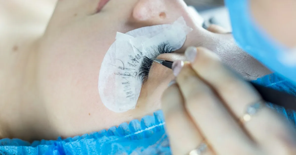 Technicienne posant des extensions de cils à la pince sur une cliente allongée, les cils inférieurs protégés par un patch